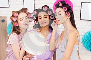 Three girls with curlers in their hair doing make up behind mirror. They are celebrating women`s day March 8.