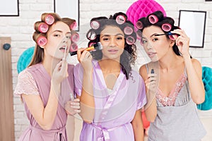 Three girls with curlers in their hair doing make up. They are celebrating women`s day March 8.