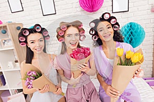 Three girls with curlers in their hair and flowers in hands. They are celebrating women`s day March 8.