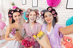 Three girls with curlers in their hair and flowers in hands taking selfie. They are celebrating women`s day March 8.