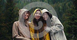 Three girls in light jackets in hoods rejoice in the coming rain and look at the camera and hug in a mountain forest