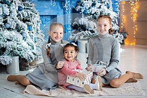 three girls are sitting near the Christmas tree. red-haired sisters, dark-skinned child unfold gifts near the fireplace