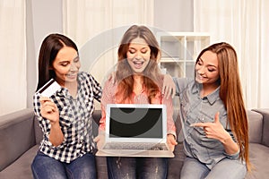 Three girls sitting on sofa and showing black screen of laptop and bank card