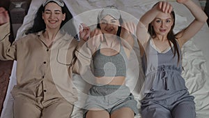 Three girls with sleep masks on their heads look at the camera while sitting on a bed in the bedroom. The girls raise