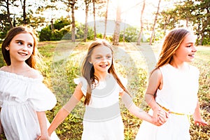 Three girls in white dresses walk in nature in the summer. Children`s pastime during the summer holidays.