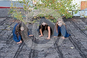 Three girls in zombie costumes crawl along the roof of an old ruined building and grimace frighteningly at the camera. Halloween photo