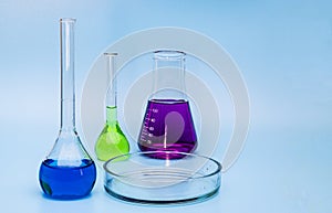 Three glass medical laboratory flasks with different multi-colored liquids and a glass bowl on a gentle blue medical background.