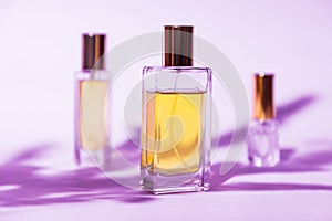 Three glass rectangular bottles of golden perfume on lilac background with sunlight and shadows of leaves