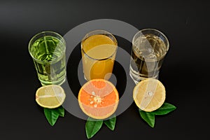 Three glasses with different juices, orange, lemon and lime on a black background, next to the leaves and pieces of ripe citrus