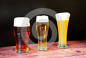 Three glasses of different types of beer with white foam stand in a row on a wooden table