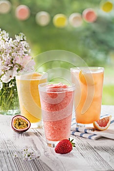 Three glasses with fresh juice from passion fruit, strawberries, red orange on a table in the garden, with ice