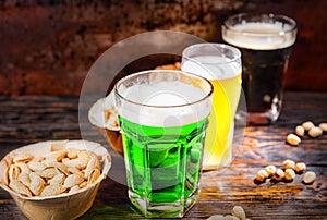 Three glasses with green, unfiltered and dark beer stand in a row near plates with snacks and scattered nuts on dark wooden desk