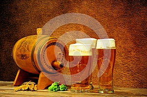Three glasses of light beer with foam are on the table next to a wooden barrel, ears of wheat and hops