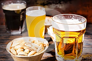 Three glasses with light, unfiltered and dark beer stand in a row near plates with different nuts on dark wooden desk