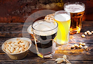 Three glasses with light, unfiltered and dark beer stand in a row near plates with snacks and scattered nuts on dark wooden desk