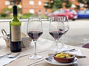 Three glasses of red wine, bottle of wine and chef`s compliment, small plate of paella served on table outdoor terrace