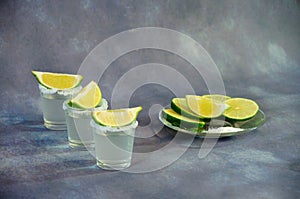Three glasses of tequila with salt at the edges and slices of lime stand in a row by the plate with salt and flat slices of citrus