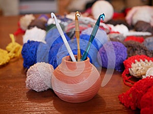 Three gold, green and white crochet hooks in a ceramic vase against a background of several multicolored balls of woolen thread on