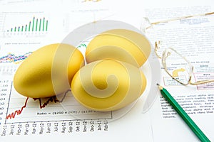 Three golden eggs with a pencil and eye glasses over business and financial summary reports.