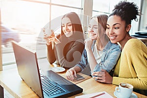 Three gourgeous best friends are in cafe. They are sitting vlose to a laptop which is on the table and loking to the photo