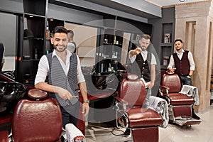 Three handsome barbers posing in barbershop near hairdresser chairs.