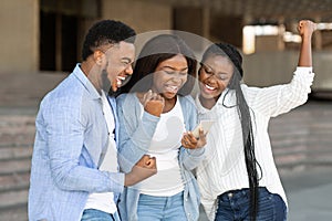 Three happy african american friends celebrating success with smartphone outdoors