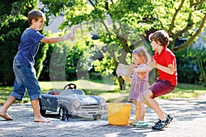 Three happy children washing big old toy car in summer garden, outdoors. Two boys and little toddler girl cleaning car