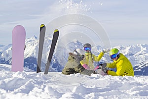 Three happy friends snowboarders and skier are having lunchtime on ski slope in sunny day in the mountains