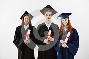 Three happy graduates smiling holding diplomas looking at camera over white background.