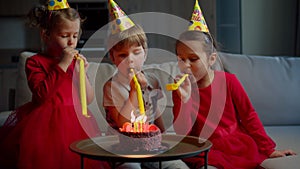 Three happy kids in birthday hats blowing birthday pipes to each other at home. Brother and sisters celebrate birthday