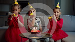 Three happy kids in birthday hats blowing birthday pipes to each other at home. Brother and sisters celebrate birthday