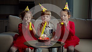 Three happy kids in birthday hats blowing birthday pipes to each other at home. Brother and sisters celebrate birthday