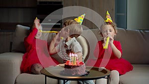 Three happy kids in birthday hats blowing birthday pipes to each other at home. Brother and sisters celebrate birthday