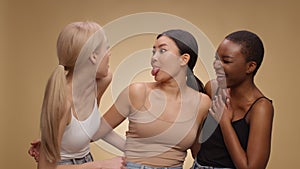 Three happy multiethnic friends laughing together, grimacing and sticking out tongues, beige background, slow motion