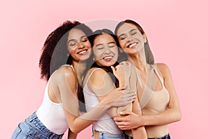 Three happy multiracial female best friends, embracing and smiling with closed eyes, posing over pink background