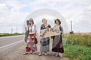 Three hippie women, wearing boho style clothes, standing on road, thumbing a ride, hitchhiking with sign Happy place on cardboard