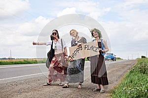 Three hippie women, wearing boho style clothes, standing on road, thumbing a ride, hitchhiking with sign Happy place on cardboard