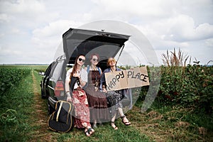 Three hippie women, wearing colorful boho style clothes, sitting on car trunk, holding Happy place sign, smiling, relaxing.