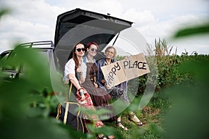 Three hippie women, wearing colorful boho style clothes, sitting on car trunk, holding Happy place sign, smiling, relaxing.