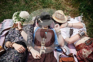 Three hippie women, wearing colorful boho style clothes and straw hats, lying, relaxing on green grass of field on sunny summer