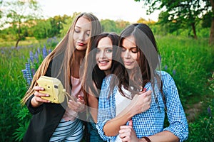Three hipsters girls blonde and brunette taking self portrait on polaroid camera and smiling outdoor. Girls having fun together