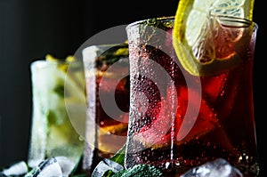 Three iced alcohol drinks on a bar with mint, lemon, juice, soda and ice.  Cocktail in different colors red and white lemonade