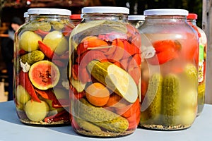 Three jars with mixed pickles on a white table for sale at a food market with pickled cucumbers, green tomatoes, red peppers, melo