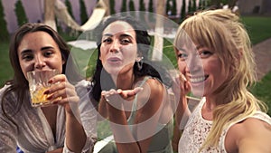 Three joyful women met in veranda cafe outdoors taking selfie photo or video on smartphone, having fun, drink