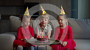 Three kids in birthday hats celebrating birthday with chocolate cake with candles at home. Brother blowing out birthdays