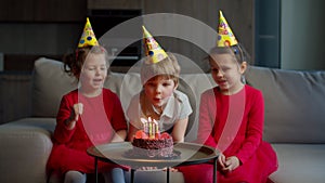 Three kids in birthday hats celebrating birthday with chocolate cake with candles at home. Brother blowing out birthdays