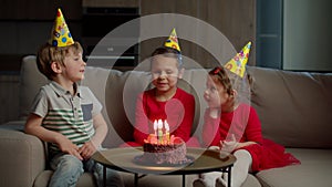 Three kids in birthday hats celebrating birthday with chocolate cake with candles at home. Girl blowing out birthdays