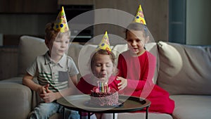 Three kids in birthday hats celebrating birthday with chocolate cake with candles at home. Girl blowing out birthdays
