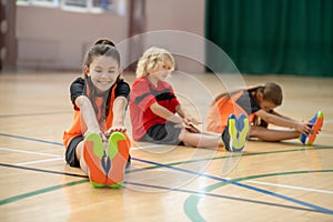 Three kids exercising in the gym and stretching froward