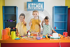 Three kids giving thumbs up at toy kitchen.